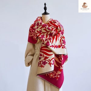 May include: A beige scarf with a floral pattern in red and fuchsia, displayed on a mannequin. The scarf has a dark red border and fringe detailing. The floral design includes stylized leaves and flowers.