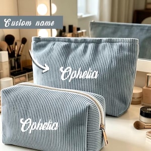 Könnte beinhalten: Zwei hellblaue Kosmetiktaschen aus Cord mit goldenen Reißverschlüssen. Die Taschen sind mit dem Namen "Ophelia" in weißer Schrift personalisiert. Die größere Tasche hat eine rechteckige Form, und die kleinere Tasche ist rechteckig.