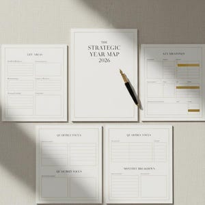 以下が含まれることがあります： 金色のアクセントとテキストが入った白い紙のプランナーのコレクション。「THE STRATEGIC YEAR MAP 2026」と書かれた中央のプランナー。金と黒のペンがプランナーの上に置かれています。他のプランナーには、生活分野、四半期ごとのフォーカス、重要なマイルストーンのセクションが含まれています。