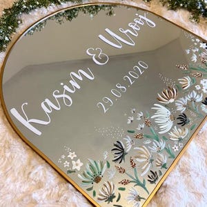 May include: A decorative mirror with a gold frame and a floral design. The mirror is inscribed with the names "Kasim & Urooj" and the date "29.08.2020" in white script. The floral design features white, green, and brown flowers.