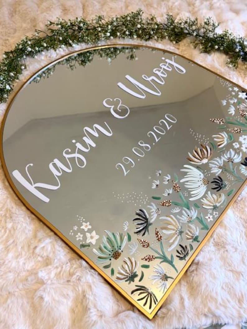 May include: A decorative mirror with a gold frame and a floral design. The mirror is inscribed with the names "Kasim & Urooj" and the date "29.08.2020" in white script. The floral design features white, green, and brown flowers.