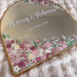 May include: A gold-framed mirror with an arched top, featuring the names "Harry & Rebecca" and the date "15th May 2026" in white script. The mirror is adorned with a floral border in shades of pink, white, and burgundy.