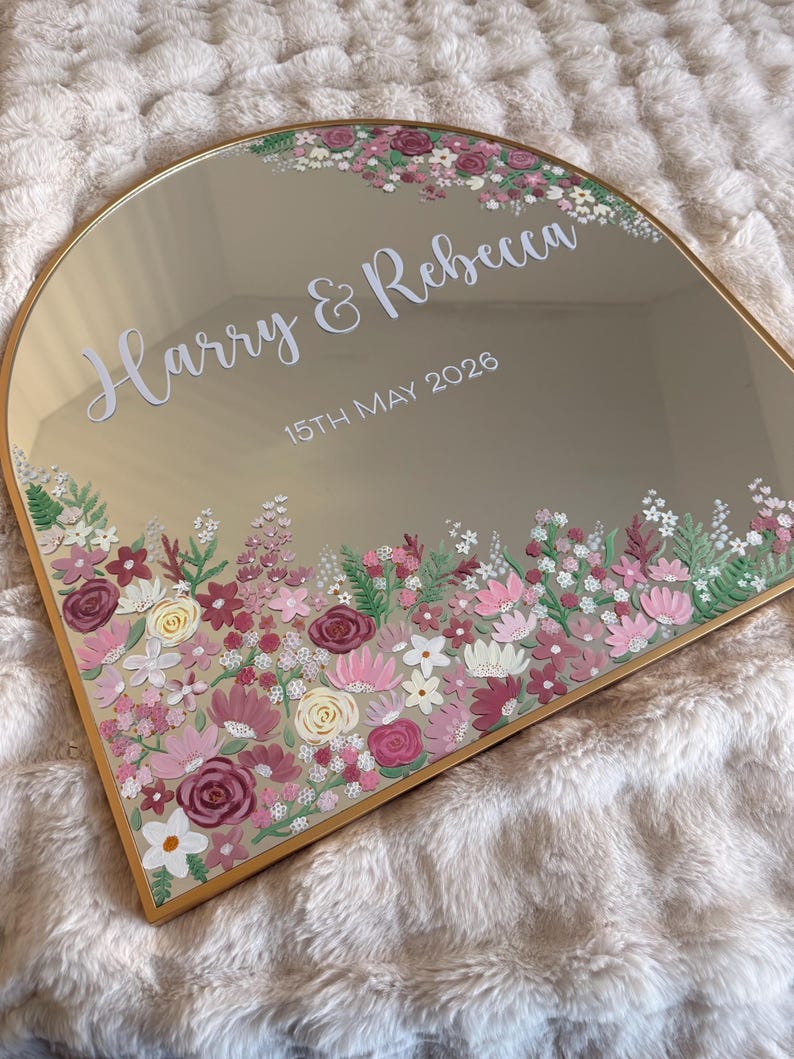 May include: A gold-framed mirror with an arched top, featuring the names "Harry & Rebecca" and the date "15th May 2026" in white script. The mirror is adorned with a floral border in shades of pink, white, and burgundy.