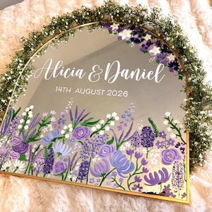 May include: A mirrored sign with a gold frame, featuring the names "Alicia & Daniel" and the date "14th August 2026". The bottom edge is adorned with a floral design in shades of purple and white, with a green floral garland around the top.