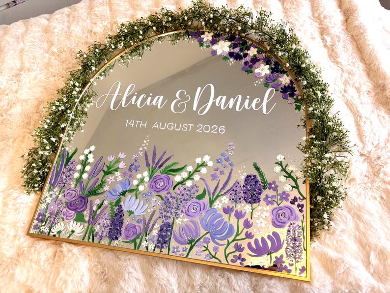 May include: A mirrored sign with a gold frame, featuring the names "Alicia & Daniel" and the date "14th August 2026". The bottom edge is adorned with a floral design in shades of purple and white, with a green floral garland around the top.