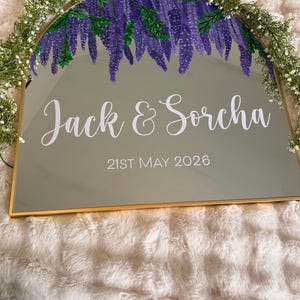 May include: A gold-framed mirror with a painted purple wisteria design at the top. The names "Jack & Sorcha" and the date "21st May 2026" are written in white script. The mirror is decorated with a garland of white flowers.