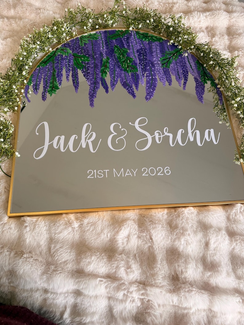 May include: A gold-framed mirror with a painted purple wisteria design at the top. The names "Jack & Sorcha" and the date "21st May 2026" are written in white script. The mirror is decorated with a garland of white flowers.
