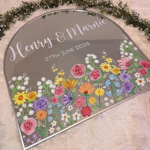 May include: A decorative, arched mirror with a silver frame. The mirror is painted with a colorful floral design along the bottom edge. The names "Henry & Marnie" and the date "27th June 2026" are written in white script.