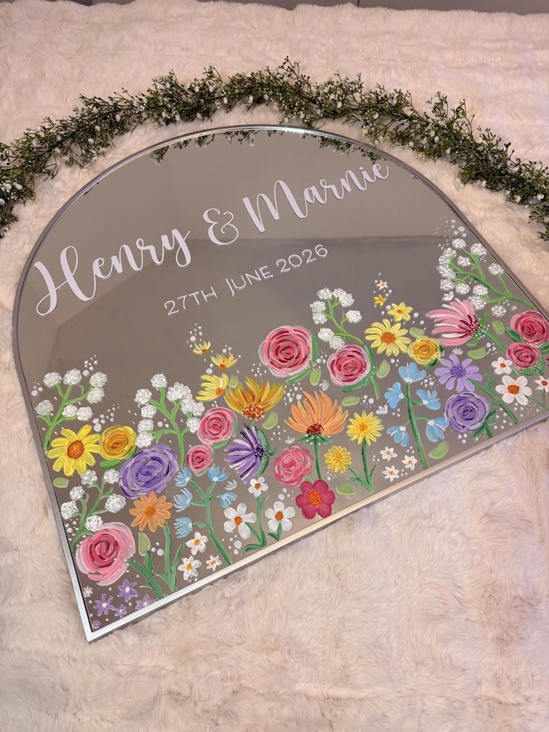 May include: A decorative, arched mirror with a silver frame. The mirror is painted with a colorful floral design along the bottom edge. The names "Henry & Marnie" and the date "27th June 2026" are written in white script.