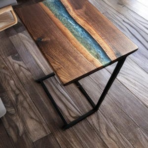 May include: A rectangular side table featuring a natural live-edge wood top with a central translucent blue and green resin inlay. The table is supported by a black metal frame with a C-shaped base, providing a stable structure for the tabletop.