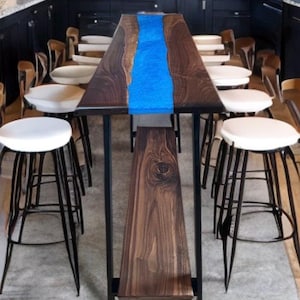 May include: A custom-made dining table with a blue epoxy river running down the center. The table is made of dark wood and has a black metal frame. White-topped stools with black metal legs surround the table.