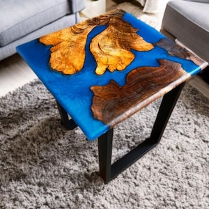 May include: Square coffee table with a blue epoxy resin top, featuring natural wood grain patterns in shades of brown and gold. The table is supported by a black metal frame, creating a modern, artistic design. The table measures approximately 24 inches square.