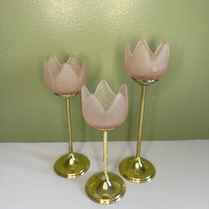 May include: Three candle holders with frosted pink glass tops shaped like tulip petals. Each holder features a slender gold-tone metal stem and a circular base. The glass elements display a delicate leaf vein texture on the exterior surface.