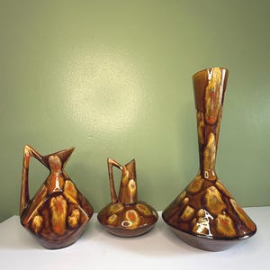 May include: A set of three ceramic vessels featuring a glossy brown glaze with mottled yellow and orange accents. The collection includes two pitchers with angular handles and one tall, slender-necked vase, all displaying a consistent drip-glaze finish.