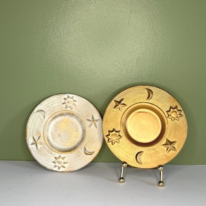 Puede incluir: Dos platos decorativos circulares con motivos celestiales. Un plato tiene un acabado en blanco desgastado con detalles dorados, mientras que el otro es de oro metálico sólido. Ambos platos muestran patrones en relieve de soles, estrellas y lunas.