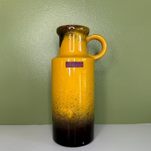 May include: A cylindrical ceramic vase featuring a vibrant yellow glaze that transitions into a deep brown at the base. The vase has a single looped handle near the top rim and a purple rectangular sticker with the text scheurich on the front.