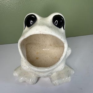 Op de afbeelding: Een keramische houder in de vorm van een kikker met een wijde, open mond. De buitenkant is gebroken wit met kleine bruine spikkels. Het object heeft twee grote, glanzend zwarte ogen bovenop de kop en vier zwemvliezen aan de onderkant.