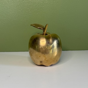 May include: A polished brass apple ornament featuring a smooth, rounded body and a single upright leaf attached to the top stem. The metallic surface reflects light, showcasing a warm golden hue and a solid, weighted appearance throughout the piece.