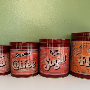 May include: A set of four cylindrical metal storage canisters in graduated sizes, featuring a deep red finish and vintage-style labels. The labels read Golden Leaf Tea, Sunshine Coffee, Brite White Sugar, and Fluffy Flour with various decorative text.