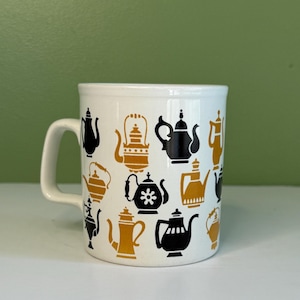 May include: A white ceramic mug featuring a repeating pattern of various teapot silhouettes in alternating black and mustard yellow colors. The mug has a simple handle on the left side and a cylindrical body with a smooth, glossy finish throughout.