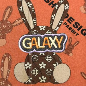 May include: A brown rabbit graphic with white flower and geometric patterns on an orange background. A central patch features the word GALAXY in yellow and orange letters with a light purple border. Additional smaller rabbit outlines are visible.