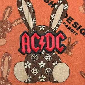 Può includere: Una toppa a forma di coniglio marrone con un motivo floreale e geometrico bianco, sovrapposta a un logo AC/DC ricamato in rosso e nero. La toppa è posta su uno sfondo arancione con un motivo ripetitivo di conigli marroni e il testo PABBIT.