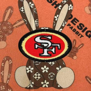 Puede incluir: Un parche bordado de forma ovalada con el logotipo de los San Francisco 49ers en rojo, blanco y negro. El parche está centrado sobre un fondo de tela marrón con un patrón de flores blancas y formas geométricas repetidas sobre una superficie naranja.