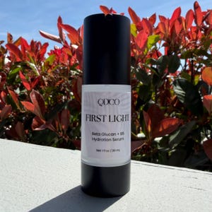 May include: A black and white bottle of QDCO First Light Beta Glucan + B5 Hydration Serum. The bottle has a white label with the product name and details. The bottle contains 1 fl oz / 30 ml of serum. The background features red and green foliage.