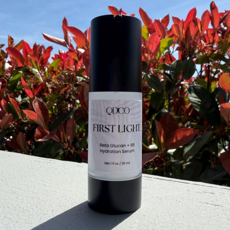 May include: A black and white bottle of QDCO First Light Beta Glucan + B5 Hydration Serum. The bottle has a white label with the product name and details. The bottle contains 1 fl oz / 30 ml of serum. The background features red and green foliage.