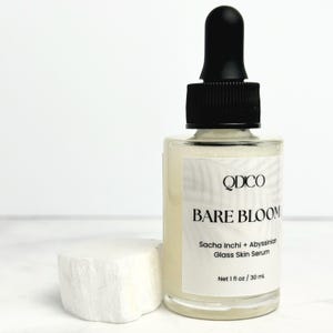 K&ouml;nnte beinhalten: Eine transparente Glasflasche mit Bare Bloom Glass Skin Serum und einer schwarzen Pipette. Die Flasche ist mit dem Markennamen QDCO und Produktdetails beschriftet. Neben der Flasche liegt ein wei&szlig;er, strukturierter Stein. Das Serum ist 30 ml.