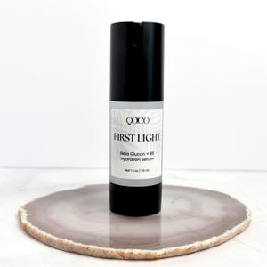May include: A black skincare serum bottle with a white label. The label reads "QDCO FIRST LIGHT Beta Glucan + B5 Hydration Serum". The bottle contains 1 fl oz / 30 ml of product. The bottle is on a decorative, round, gray and white stone.