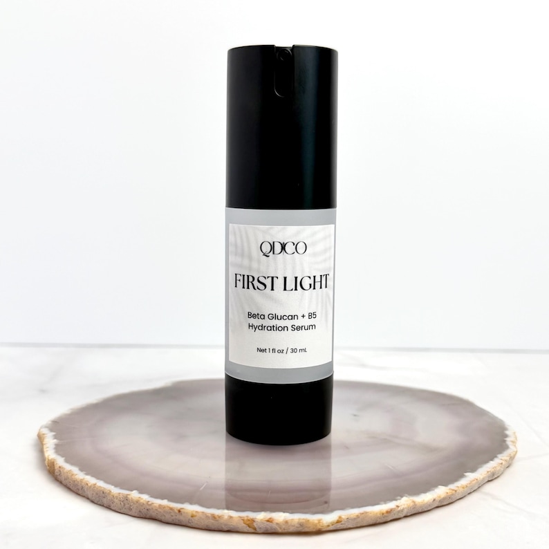 May include: A black skincare serum bottle with a white label. The label reads "QDCO FIRST LIGHT Beta Glucan + B5 Hydration Serum". The bottle contains 1 fl oz / 30 ml of product. The bottle is on a decorative, round, gray and white stone.