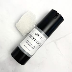 May include: A black and white bottle of "FIRST LIGHT" hydration serum with a white label. The label includes the brand name "QDCO" and the text "Beta Glucan + B5 Hydration Serum". A white, textured rock is next to the bottle. The bottle contains 1 fl oz / 30 ml.
