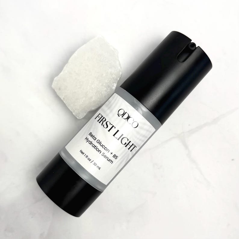 May include: A black and white bottle of "FIRST LIGHT" hydration serum with a white label. The label includes the brand name "QDCO" and the text "Beta Glucan + B5 Hydration Serum". A white, textured rock is next to the bottle. The bottle contains 1 fl oz / 30 ml.