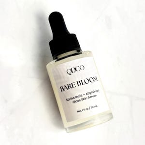 K&ouml;nnte beinhalten: Eine klare Glas-Serumflasche mit schwarzer Pipette. Auf dem Etikett steht "QDCO BARE BLOOM" mit Text darunter, der darauf hinweist, dass das Serum Sacha Inchi und Abyssinian-&Ouml;l enth&auml;lt. Die Flasche enth&auml;lt 30 ml Serum.