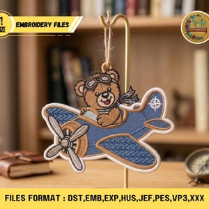Könnte beinhalten: Ein gestickter Aufnäher mit einem braunen Teddybären mit Fliegerbrille und Schal, der ein blaues Flugzeug steuert. Das Flugzeug hat einen Propeller und ein weißes Sternemblem. Der Text "EMBROIDERY FILES" wird in einem gelben Banner angezeigt. Der Aufnäher hängt an einem goldenen Ständer.