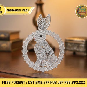 Op de afbeelding: Een gedetailleerd wit borduurbestand met een konijn in een bloemenkrans. De afbeelding bevat de tekst "EMBROIDERY FILES" en "FILES FORMAT: DST, EMB, EXP, HUS, JEF, PES, VP3,XXX". Het konijnontwerp heeft ingewikkelde patronen.
