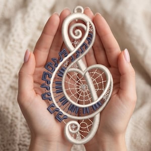 May include: Embroidered treble clef ornament with a dreamcatcher design. The white ornament features a heart-shaped dreamcatcher, piano keys, and musical notes in navy blue. Decorative stars are also embroidered on the ornament.