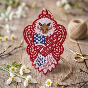 Può includere: Un ornamento patriottico con un design di aquila ricamata e la bandiera americana. L'ornamento è rosso, bianco e blu, con dettagli intricati. È appeso con uno spago naturale. Lo sfondo include fiori e una superficie di legno.