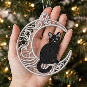 Peut inclure: Une décoration en forme de croissant de lune brodé argenté avec un chat noir assis sur la courbe inférieure. Le motif de la lune présente des volutes complexes et des découpes en forme d'étoile, surmonté d'une boucle florale pour suspendre.
