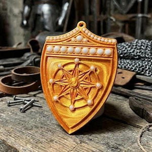 May include: A shield-shaped ornament in a metallic gold finish, featuring a central compass rose design and a border accented with small white spherical beads. The piece includes a small loop at the top for hanging and has a textured surface.