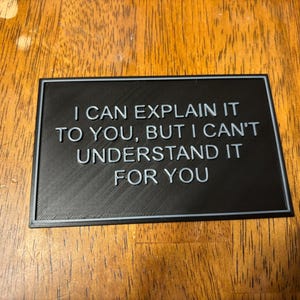 May include: A black rectangular sign with the phrase "I CAN EXPLAIN IT TO YOU, BUT I CAN'T UNDERSTAND IT FOR YOU" in light gray lettering. The sign has a thin, light gray border and is set on a wooden surface.