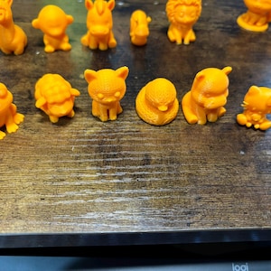 May include: A collection of small, orange animal figurines, including a giraffe, frog, cat, and lion, are arranged on a wooden surface. The figurines are detailed and appear to be 3D-printed, showcasing various animal forms.
