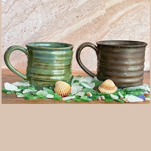 May include: Two handmade ceramic mugs with ribbed detailing. One mug is green, the other is brown. The mugs have large handles and sit on a bed of green glass pieces and seashells. The mugs are likely for coffee or tea.