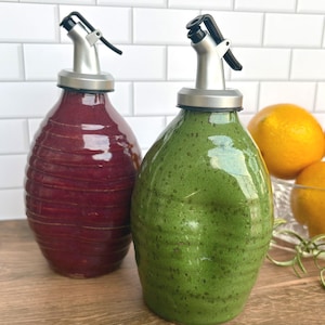 May include: Two ceramic oil and vinegar bottles with stainless steel spouts. One bottle is a deep burgundy color with horizontal ridges, and the other is a textured green. Oranges are visible in the background.