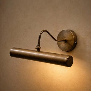 Puede incluir: Una lámpara de latón para cuadros. La lámpara tiene una carcasa de luz cilíndrica, un brazo curvo y un soporte de pared circular. La luz está montada en una pared de color neutro y está encendida.