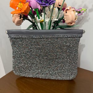 Puede incluir: Un clutch de malla plateada con una parte superior de cuero gris oscuro. El clutch es rectangular y tiene una textura tejida. El fondo presenta un colorido arreglo de flores artificiales.