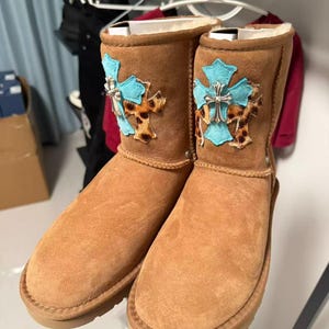 May include: A pair of tan suede ankle boots featuring a light blue cross patch layered over a leopard print fabric patch on the outer side. Each boot is adorned with a metallic silver cross emblem centered on the light blue patch, with a plush lining.