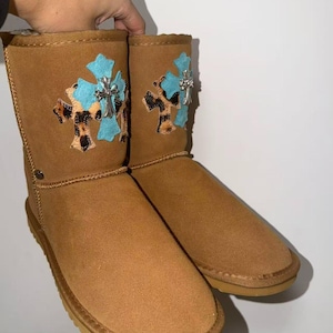 May include: Brown suede boots with a light brown sole. Each boot features a turquoise cross with a silver embellishment and a leopard print design. The boots are held up by a hand.
