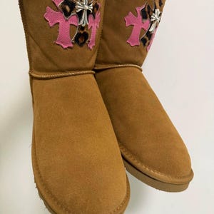 May include: A pair of tan suede boots featuring a decorative patch on the upper shaft. The patch consists of a pink cross layered with a leopard print cross and a silver-toned metal cross emblem centered on top of the layered cross design.